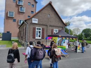 Eine Gruppe von Menschen steht vor einem Pavillon auf einem asphaltierten Platz. Im Pavillon stehen Tische mit Flyern und Stickern. Der Pavillon ist mit bunten Fahnen aus der LSBTI Community geschmückt. Um den Pavillon herum stehen Liegestühle, ein Glücksrad und ein Wurfspiel. Im Hintergrund ist ein Haus, auf dem ein "ASTA" Schild steht. Es ist der Campus der Hochschule Rhein Waal.