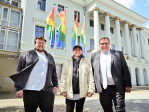 Drei Menschen stehen lächelnd vor einem hohen Gebäude, dem Radhaus. Im Hintergrund sind vier Fahnenmasten mit Regenbogenflaggen gehisst. ZU sehen sind Jan-Eric vom together krefeld, Pia Günther von der Aidshilfr und SCHLAU Krefeld gemeinsam mit dem Oberbürgermeistwer Frank Meyer..